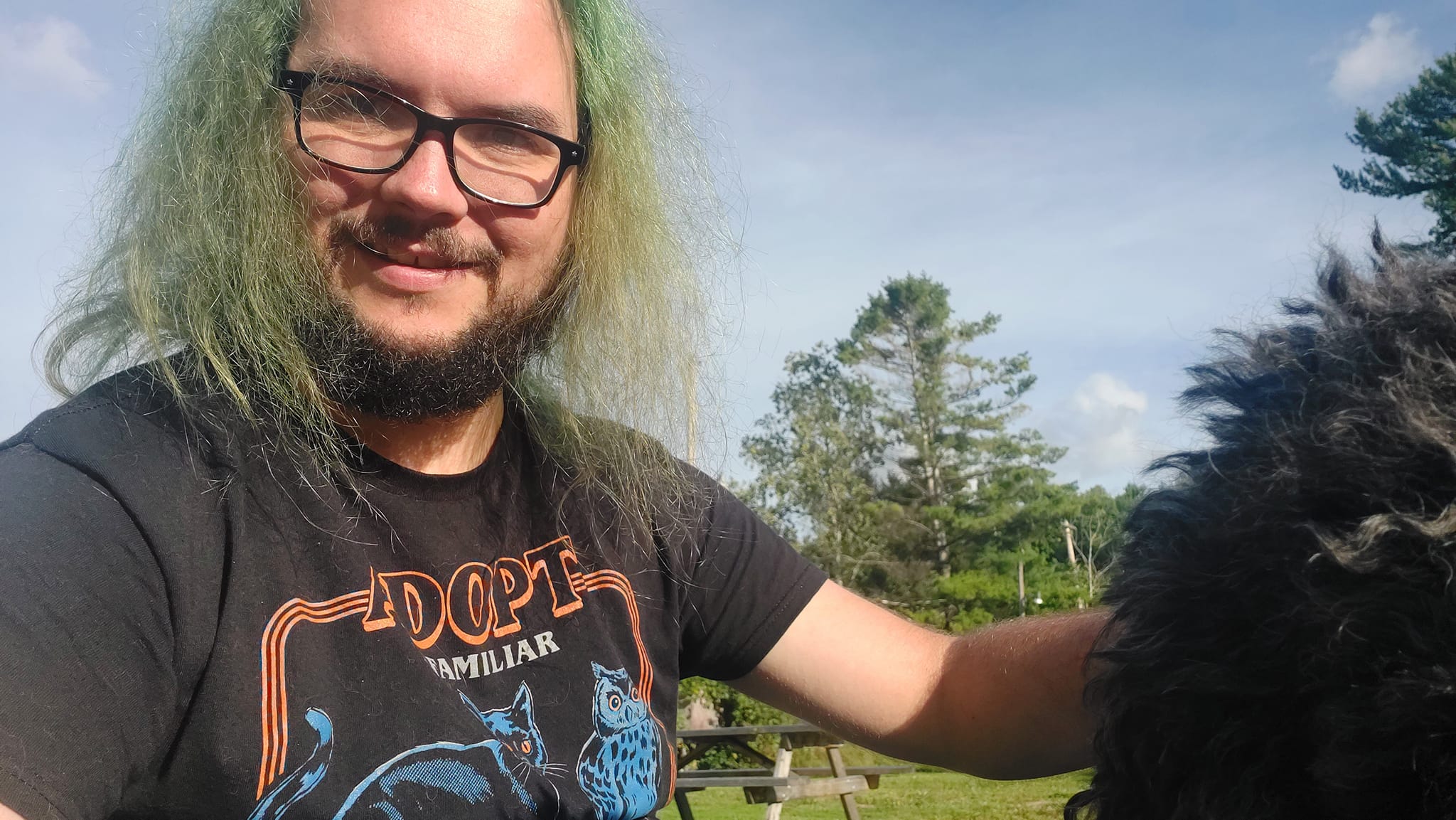 A man with green hair and a beard is outside with a black standard poodle out of frame, Photo 1